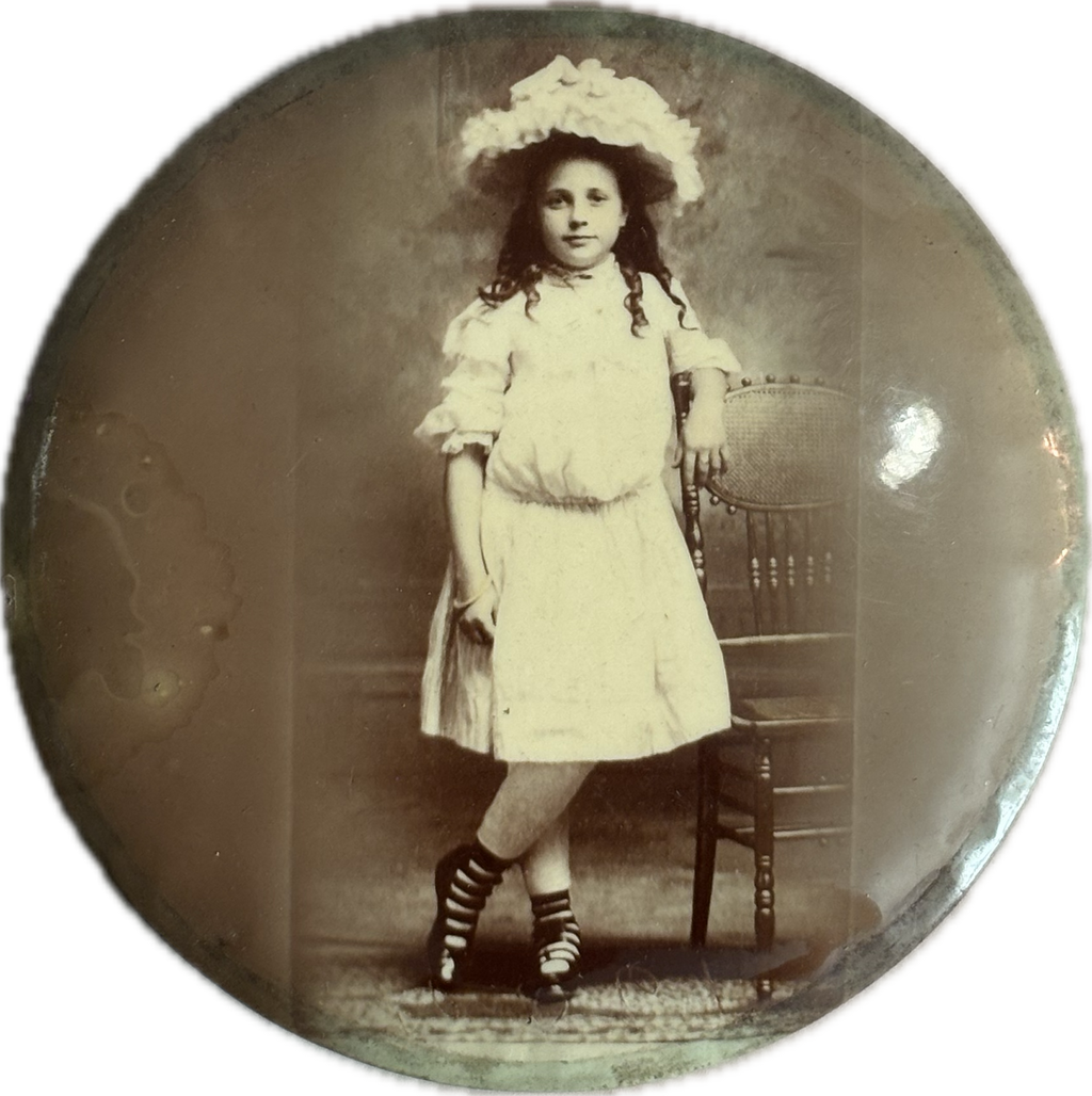 Vintage photograph of a young girl in a white dress and hat, standing on a chair.